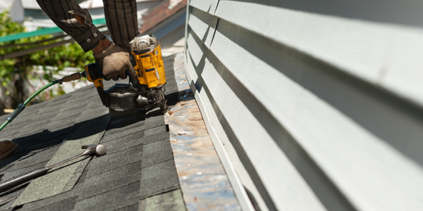 hands using a nail gun to install asphalt shingles,  you can see house siding on the right